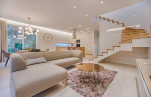 Interno casa vacanze con soggiorno e cucina open-space, divano beige, tavolino rotondo in metallo, scale in legno e pareti bianche illuminate. - Photo by Vecislavas Popa/pexels.com