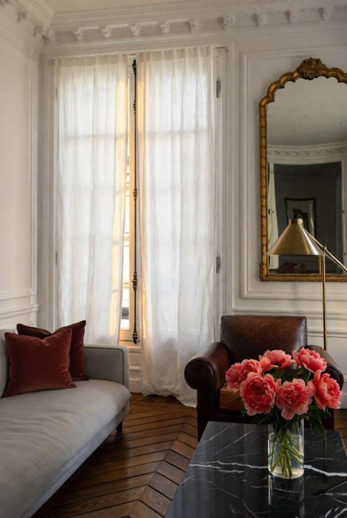 Arredare in stile parigino con angolo elegante: poltrona cuoio, divano grigio, parquet a spina di pesce e specchiera dorata classica.