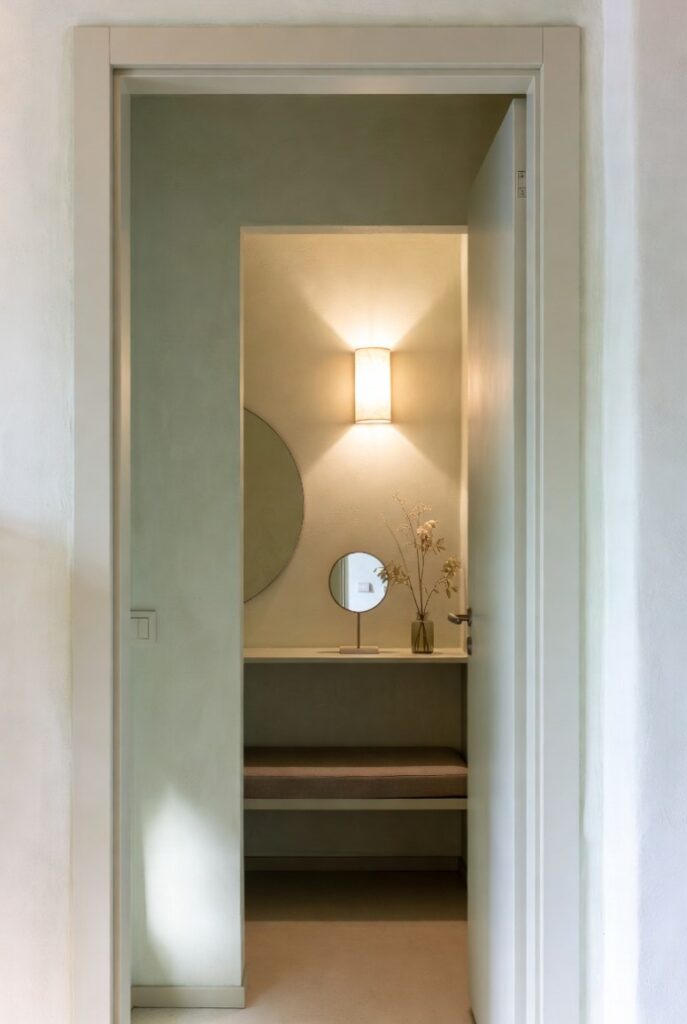 Effetto scatola ingresso piccolo con pareti verde chiaro uniforme, specchio rotondo, applique luminosa e panca, design minimal luminoso 