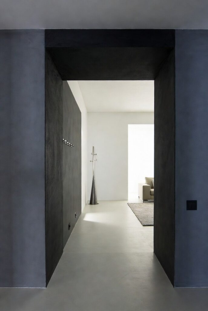Effetto scatola ingresso grigio antracite con confine netto apertura soggiorno bianco, appendiabraccio, luce naturale e pavimento cemento divisione spazi