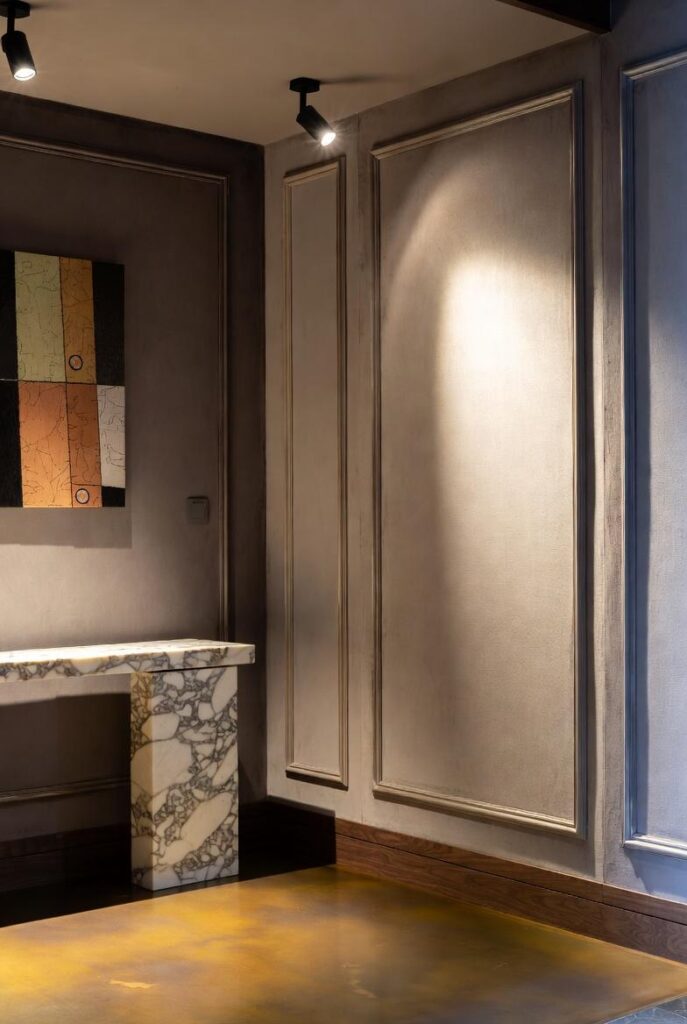 Effetto scatola ingresso grigio con faretti orientabili luce calda su quadro astratto, consolle marmo boiserie, pavimento oro per combo illuminazione materiali