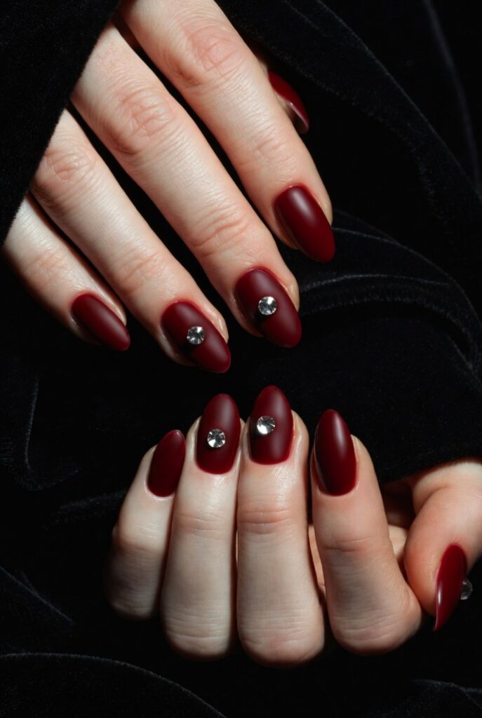 Mani close-up unghie vinaccia Valentino matte scura con strass centrale, sfondo nero velluto – idee rosso Valentino storia per inverno. 