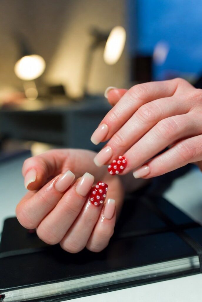 Mani close-up manicure minimal: unghia anulare rossa Valentino con mini-perle bianche, altre nude su sfondo ufficio – idee rosso Valentino storia eleganti
