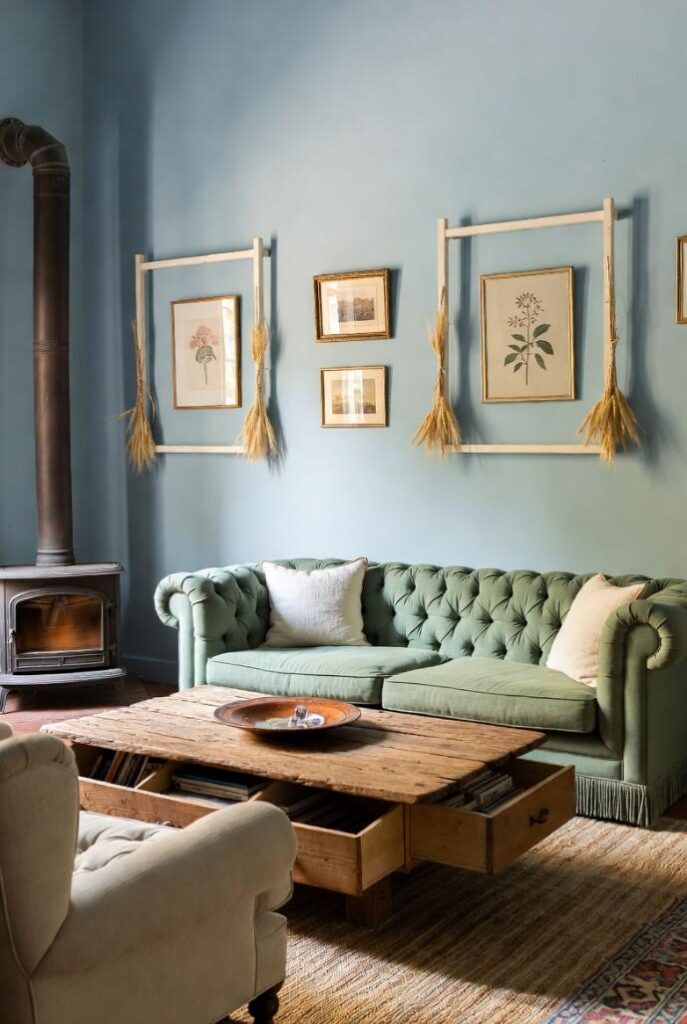 Soggiorno cozy in stile provenzale shabby chic con divano Chesterfield bouclé verde salvia, poltrone bergère beige, tavolino legno grezzo con cassetti, pareti azzurro polvere decorate con stampe botaniche e ghirlande spighe, tappeto juta naturale, stufa ghisa accesa, luce crepuscolare calda