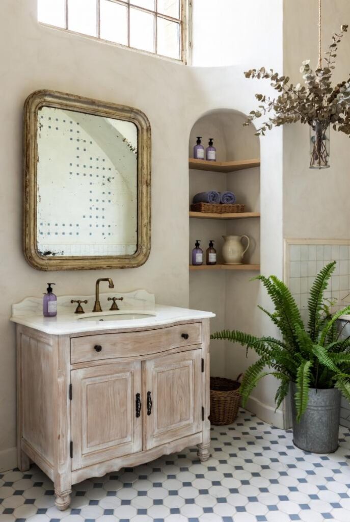 Bagno spa in stile provenzale shabby chic con mobile lavabo decapato, specchio ovale anticato, piastrelle esagonali bianco-azzurro, pianta felce e eucalipto appeso, luce naturale soffusa, colori pastello tenui.