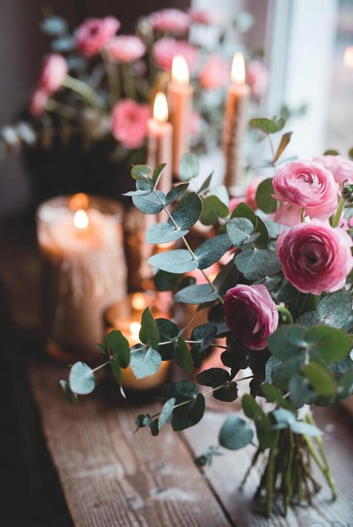 Immagine di decorazioni floreali con eucalipto rosa tinto, ranuncoli e candele accese su tavolo rustico, per un'atmosfera calda e naturale.