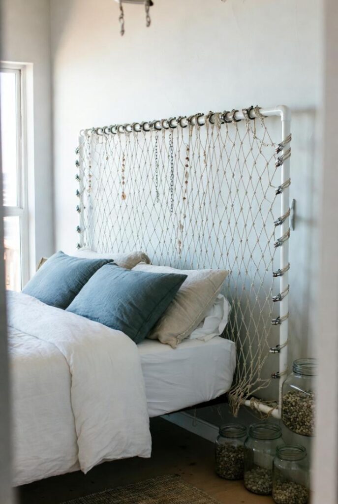 Cosa usare come testata del letto: rete da pesca bianca tesa su tubi PVC struttura, letto matrimoniale lenzuola bianche cuscini azzurri beige, barattoli vetro con conchiglie, camera boho costiera