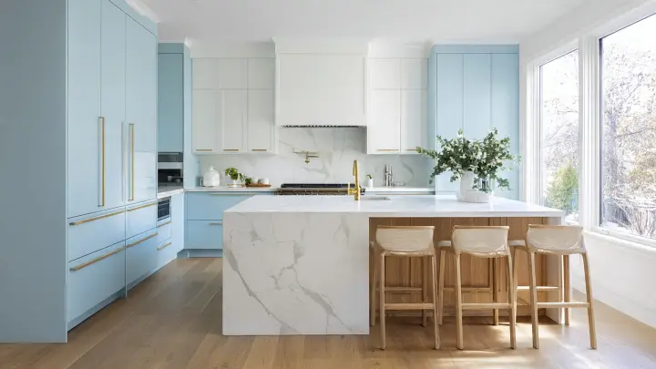 Cucina moderna celeste e bianca con grande isola in marmo bianco, colonne azzurro cielo con maniglie dorate, sgabelli in legno e rattan e pavimento in rovere chiaro