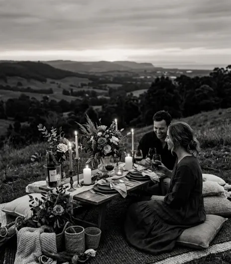 Picnic al tramonto su collina con vista valle, coppia seduta a tavola bassa elegante con candele e fiori, atmosfera romantica lusso, fotografia bianco e nero ultra realistica