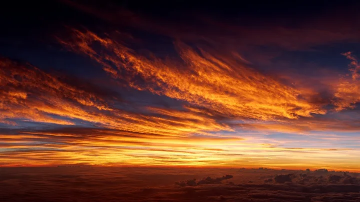 Tramonto spettacolare sull'orizzonte terrestre con cielo intenso arancione, rosso e dorato, luce solare che attraversa l'atmosfera densa, nuvole drammatiche illuminate da luce calda, fotografia paesaggistica fotorealistica stile National Geographic, illuminazione cinematografica, HDR ultra dettagliato, 16:9 senza persone né testo