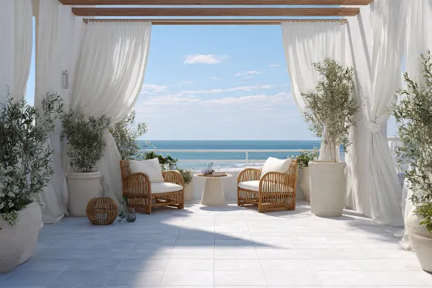 Terrazza luminosa con pavimento chiaro, tende bianche leggere, due poltrone in rattan con cuscini bianchi, vasi con ulivi e piante, tavolo basso centrale, vista aperta sul mare azzurro