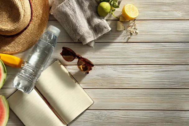 Composizione dall'alto su tavolo in legno chiaro con cappello di paglia, bottiglia d'acqua trasparente, occhiali da sole marroni, quaderno aperto a pagine bianche, asciugamano beige, lime intero, mezzo limone, fette di anguria e tubetto di crema solare gialla