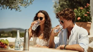 Coppia giovane beve acqua da bottiglie trasparenti seduta a un tavolo esterno sotto un ulivo, sul tavolo borraccia in acciaio, bottiglie di vetro e ciotola con uva e fragole, sullo sfondo colline e cielo azzurro estivo