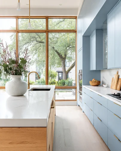 Cucina moderna color celeste polvere con mobili laccati, isola in legno chiaro con top bianco, rubinetto e maniglie in ottone, ampia vetrata con infissi in legno