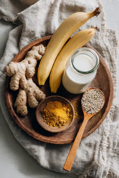 Ingredienti per frullato anti gonfiore su vassoio in legno: banane mature, radice di zenzero fresco, curcuma in polvere, semi di chia e latte vegetale