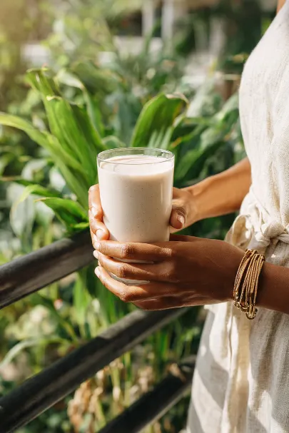 Donna che tiene con entrambe le mani un bicchiere di frullato cremoso chiaro, all'aperto tra piante verdi
