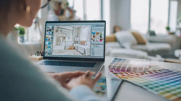 Giovane designer che lavora al laptop con software di interior design e planimetria 3D visibile sullo schermo. Scrivania moderna minimal con tablet e campioni di palette colori, stanza luminosa e accogliente. Stile fotografia realistica con luce naturale soffusa, profondità di campo ridotta, estetica Pinterest. Formato 16:9.