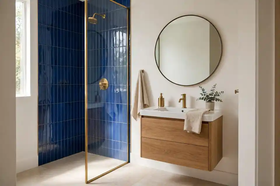 Piccolo bagno con parete doccia in piastrelle blu cobalto e mobile in rovere Piccolo bagno con parete doccia rivestita in piastrelle blu cobalto rettangolari posate in verticale, mobile lavabo sospeso in rovere naturale, lavabo bianco e specchio rotondo con bordo nero