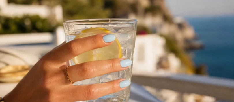 Primo piano di una mano femminile con manicure a mandorla colore unghie celeste pastello, mentre tiene un bicchiere d'acqua ghiacciata con limone in un'ambientazione estiva mediterranea al tramonto.