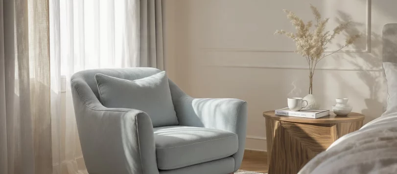 Angolo relax in una camera matrimoniale celeste e legno con poltrona azzurra, tavolino in noce e luce naturale che filtra dalle tende, stile moderno ed elegante.