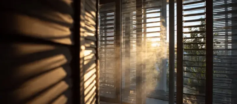 Interno di un appartamento moderno in piena estate con finestre chiuse, tapparelle semiaperte, raggi di sole che entrano e aria calda visibile con effetto shimmer. Atmosfera afosa e realistica, stile fotografia editoriale di design con colori naturali e luce estiva italiana.