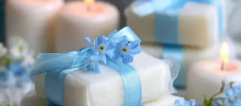 Saponette profumate fatte a mano per bomboniere battesimo, confezionate con organza bianca e nastro celeste, decorate con fiorellini azzurri su sfondo di candele accese