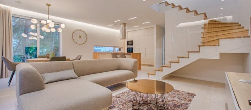 Interno casa vacanze con soggiorno e cucina open-space, divano beige, tavolino rotondo in metallo, scale in legno e pareti bianche illuminate. - Photo by Vecislavas Popa/pexels.com